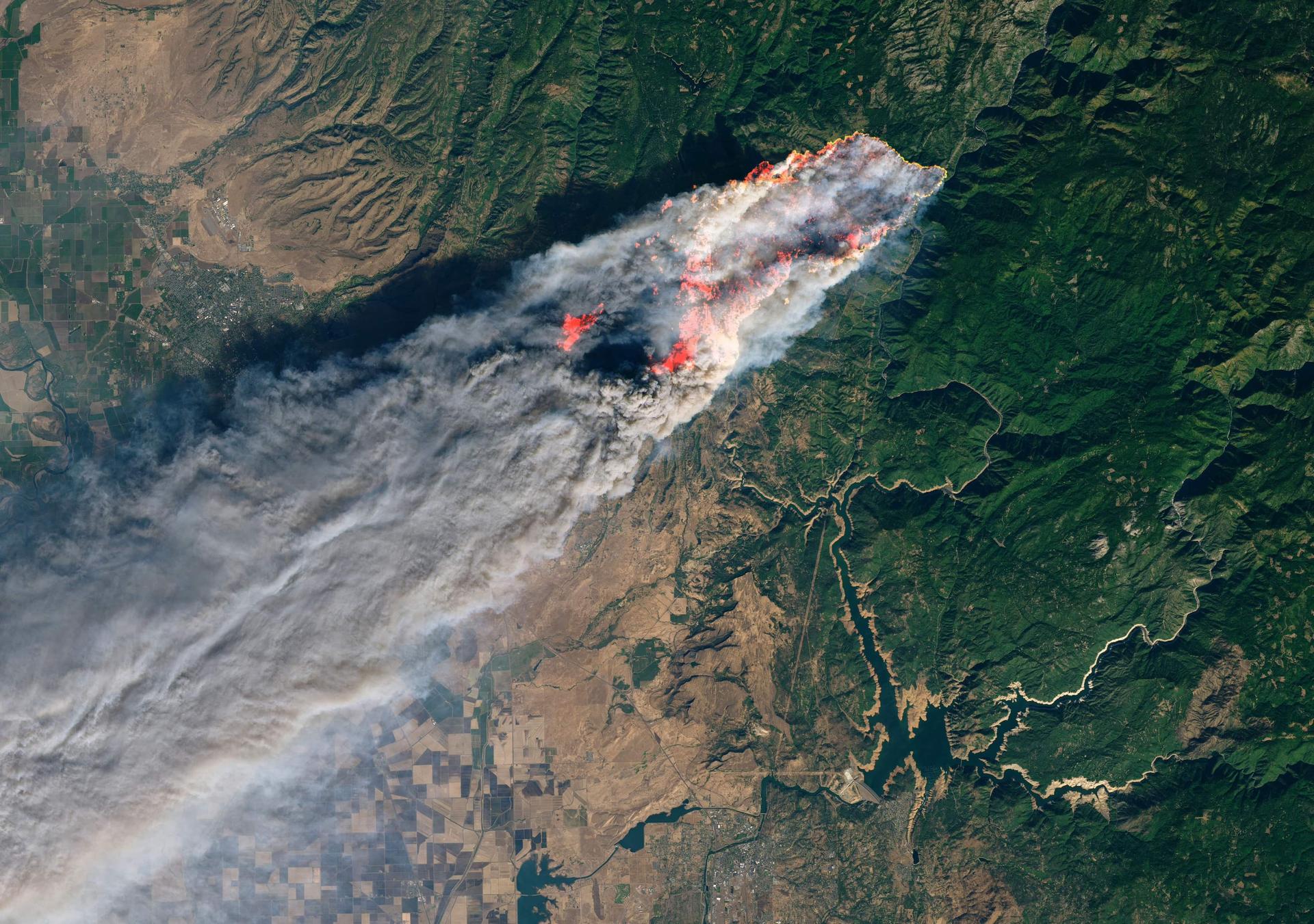 Satellite image showing a large plume of smoke from an ongoing wildfire spreading across a vast landscap/homee with visible patches of burning areas.