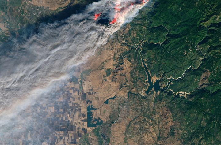 Satellite image showing a large plume of smoke from an ongoing wildfire spreading across a vast landscap/homee with visible patches of burning areas.